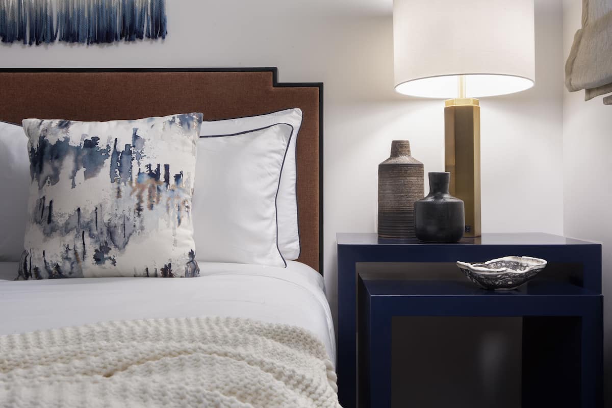 A neatly made bed with a white comforter and two pillows, a bedside table with a lamp, and a decorative bowl.