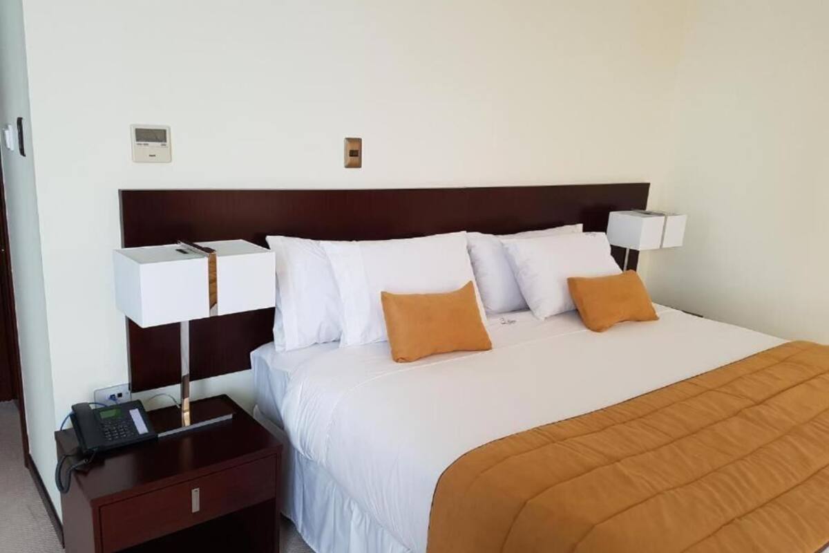 Une chambre d’hôtel avec un grand lit, deux tables de chevet, une petite table, un thermostat fixé au mur et un téléphone.