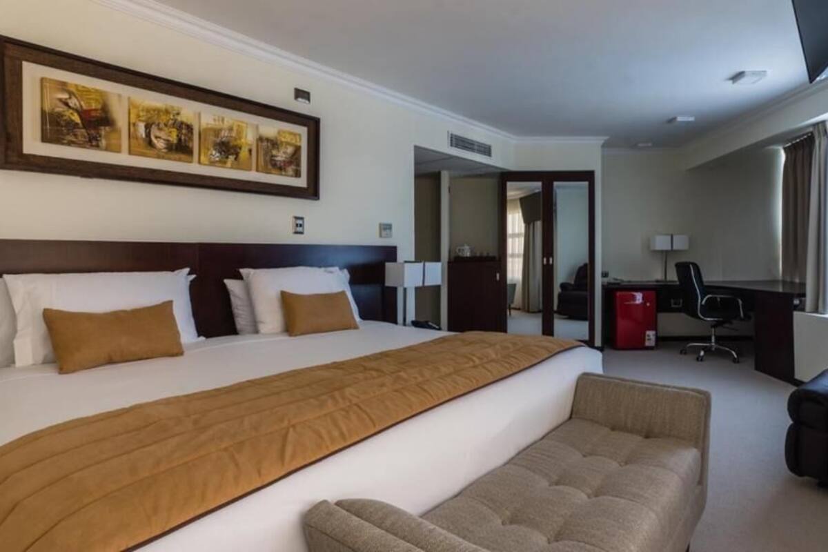 Une chambre d’hôtel avec un grand lit, un bureau, une chaise et un canapé. Il y a un tableau au mur et une fenêtre avec des rideaux.