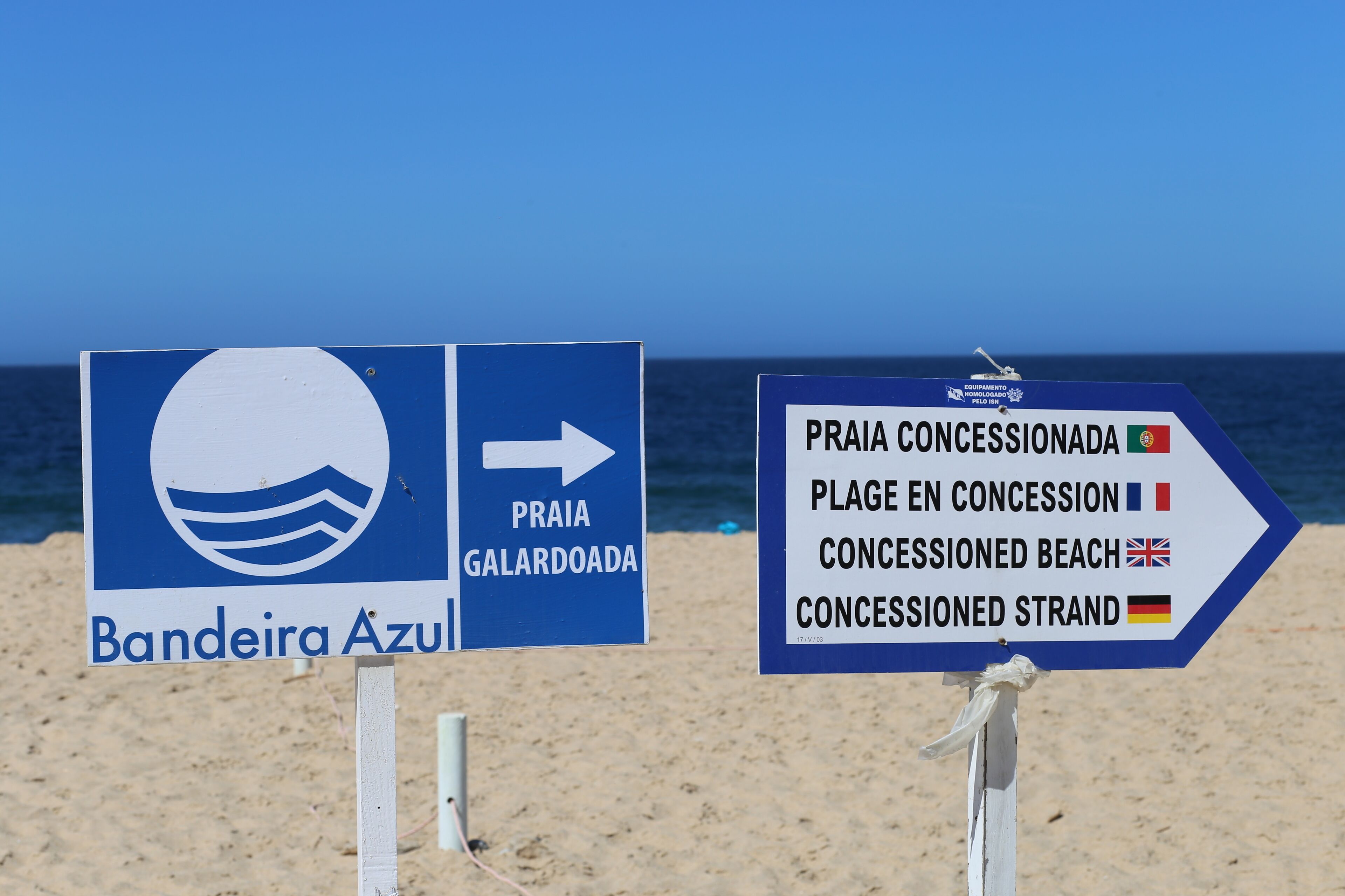 Twee richtingaanwijzers op een strand, waarvan één naar Praia Galardoada wijst en de ander naar een strandgedeelte met horeca.