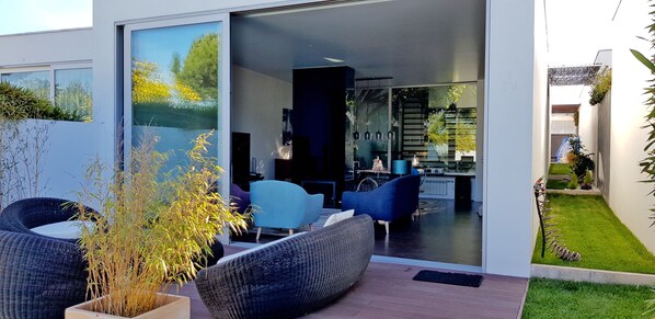 A modern house with a glass door leading to a living area with blue chairs and a dining area.