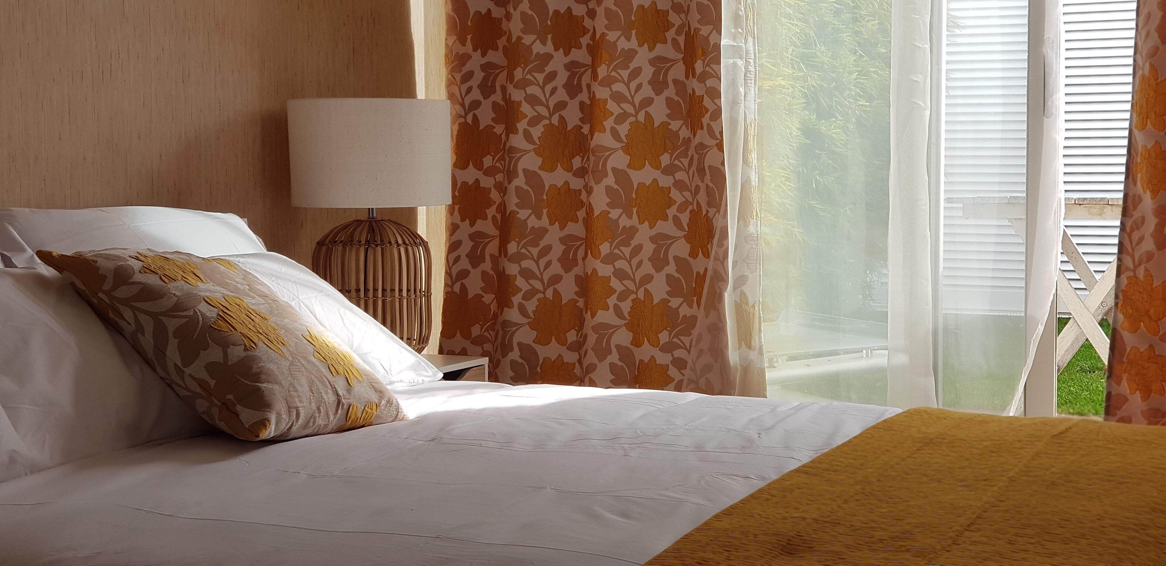 A neatly made bed with white linens and a yellow throw blanket, a bedside lamp, and a window with floral curtains.