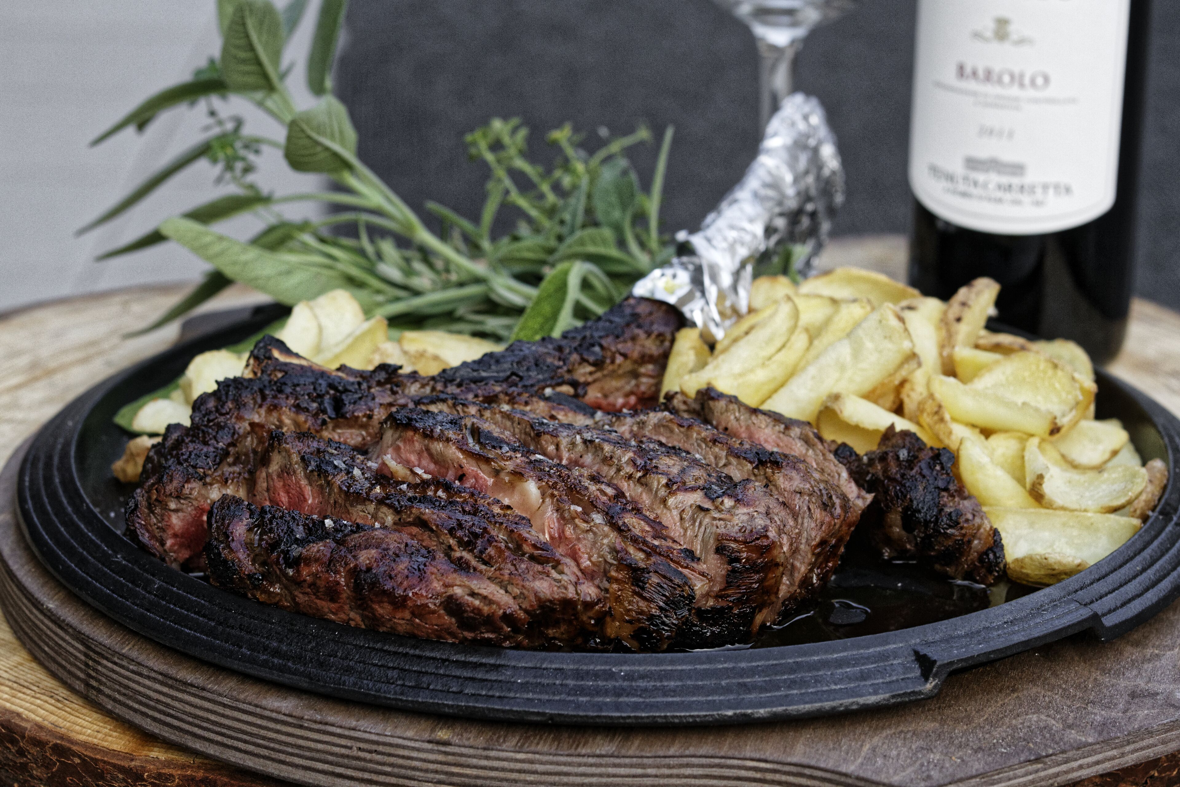 A plate with grilled steak and fries, a bottle of Barolo wine, and some herbs.
