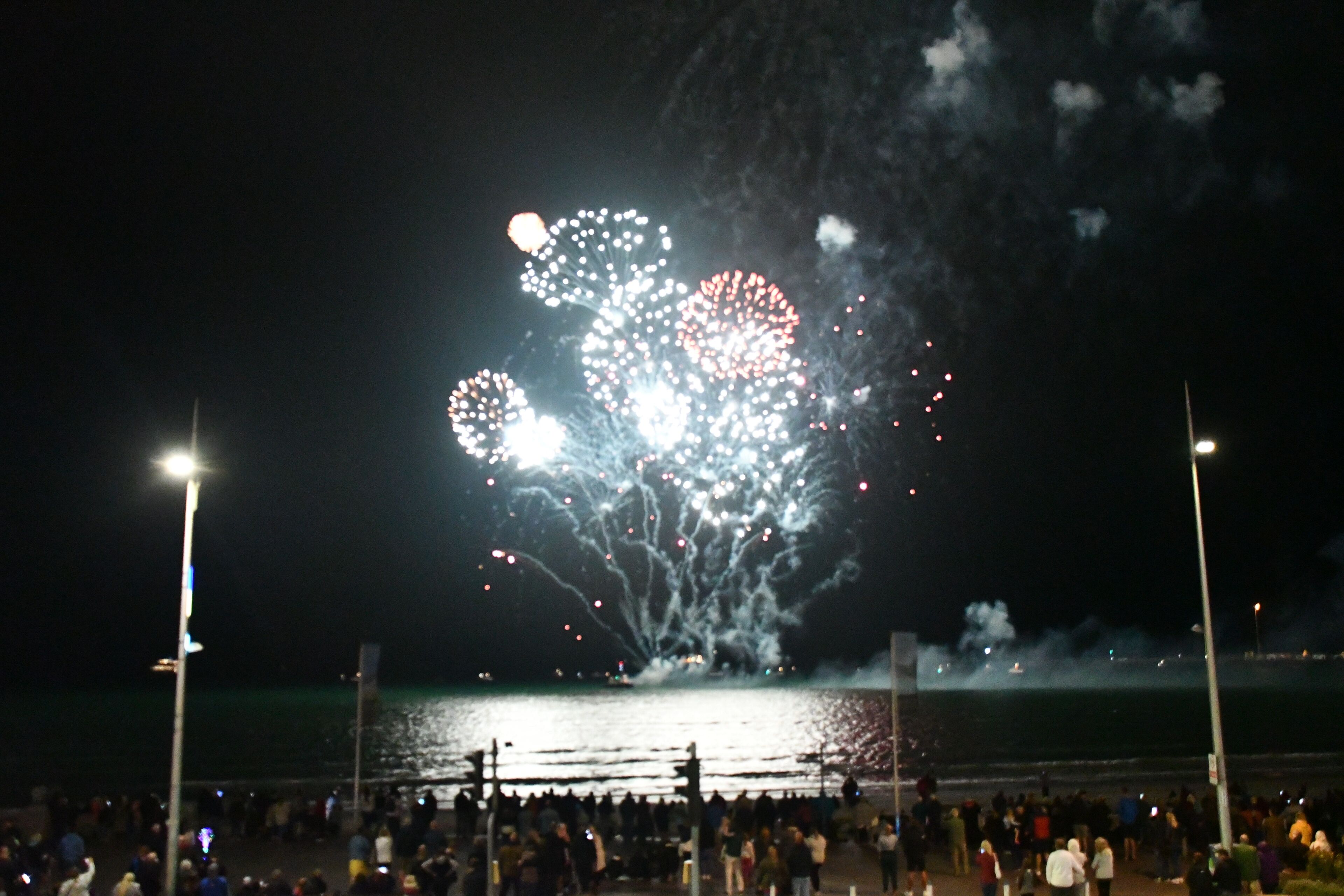 A nighttime scene with fireworks over a body of water, a crowd of people, and street lamps.