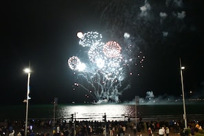 A nighttime scene with fireworks over a body of water, a crowd of people, and street lamps. - No 98 Boutique Hotel (Weymouth)