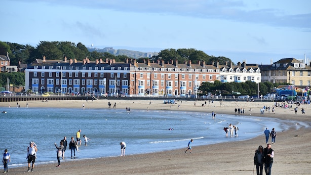 Een strand met mensen die langs de kust wandelen, een rij kleurrijke terrashuizen op de achtergrond, en een heldere lucht.