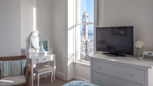 Een slaapkamer met een televisie, een dressoir, een stoel, een kaptafel met spiegel en een raam met uitzicht op een klokkentoren.