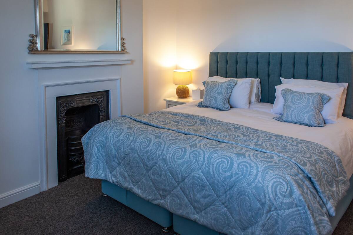 A bedroom with a bed, a nightstand, a lamp, and a fireplace.