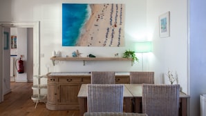 Een eetruimte met rieten stoelen, een houten tafel en een schilderij met een strandthema erboven.