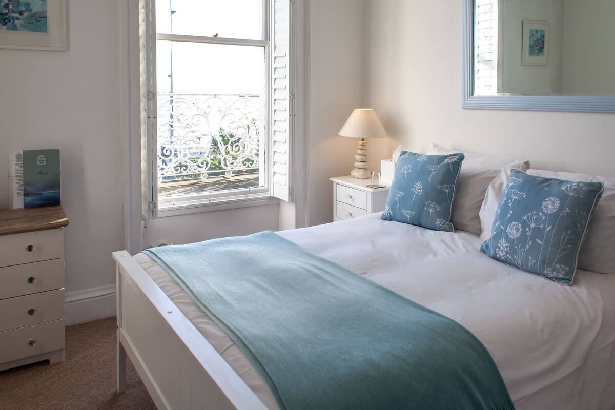 A neatly arranged bedroom with a bed, a bedside table with a lamp, a window with a view, and a dresser.