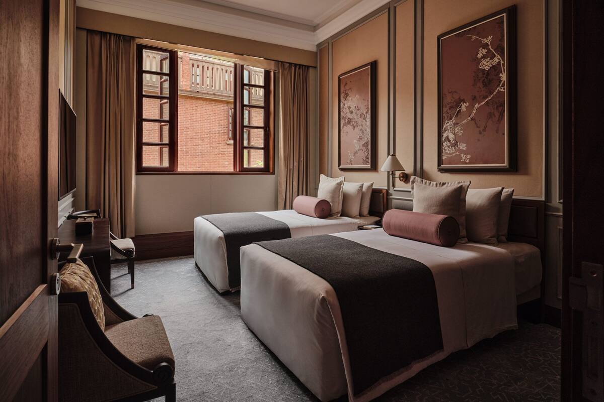 A hotel room with two beds, a desk, and a chair. There are large windows with curtains, a lamp, and artwork on the walls.
