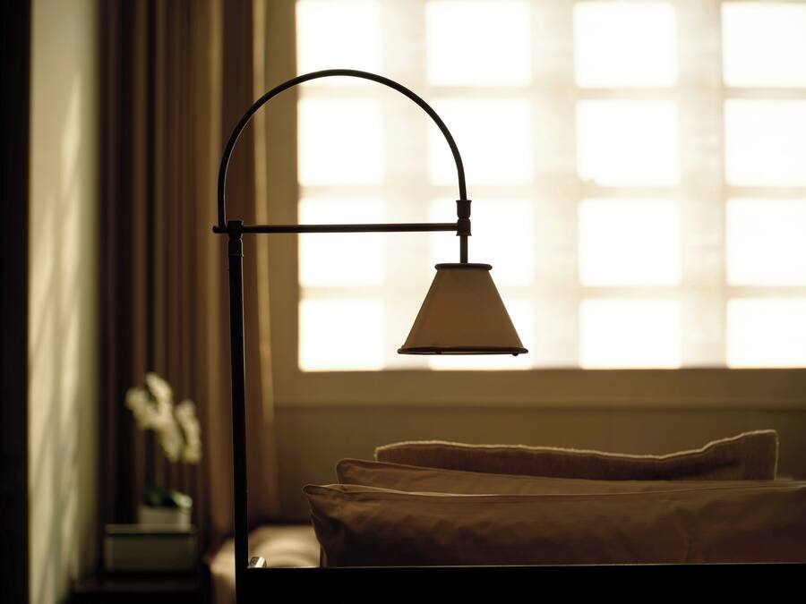 Una cama con cubre blanco, una mesita de noche con lámpara y una ventana con persianas.