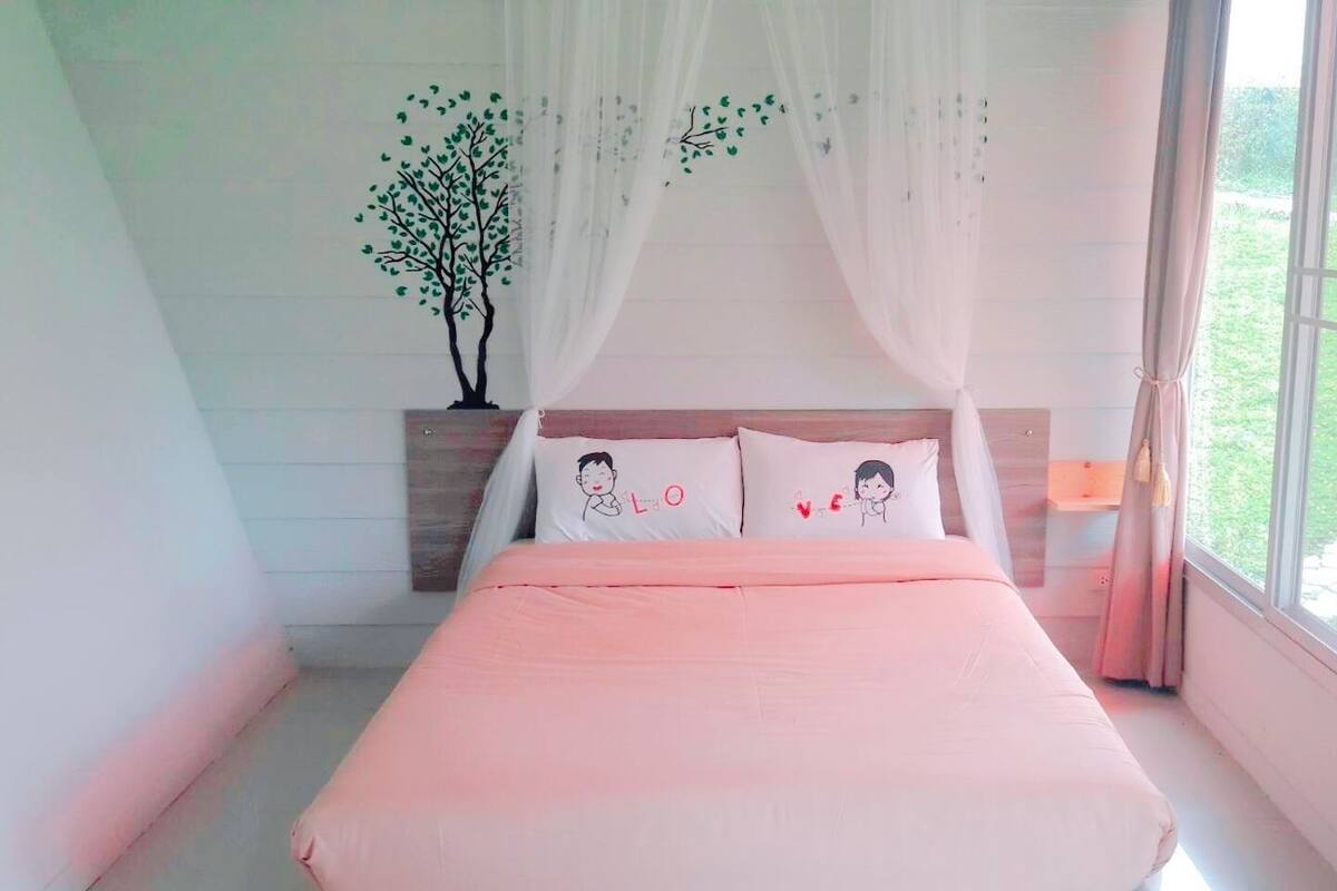 Quarto com cama, mesa de cabeceira, janela com cortinas e um adesivo de árvore na parede.