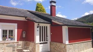 Ein rotes und weißes Haus mit Steinuntergrund, Schornstein und einer kleinen Terrasse mit Tisch und Stühlen.