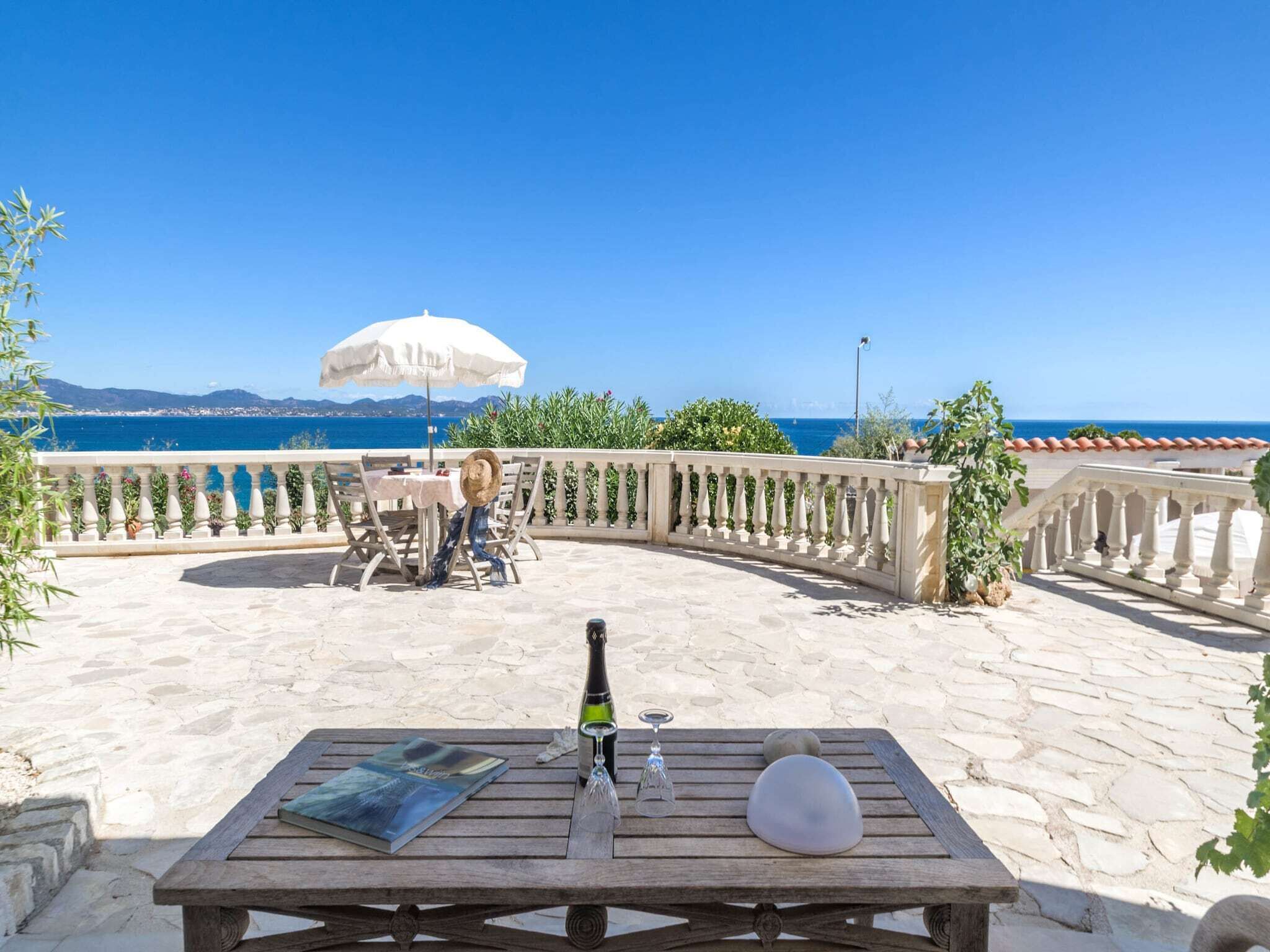 A stone patio with a wooden table, a bottle of champagne, and a glass. There is a white umbrella and a view of the sea in the background.