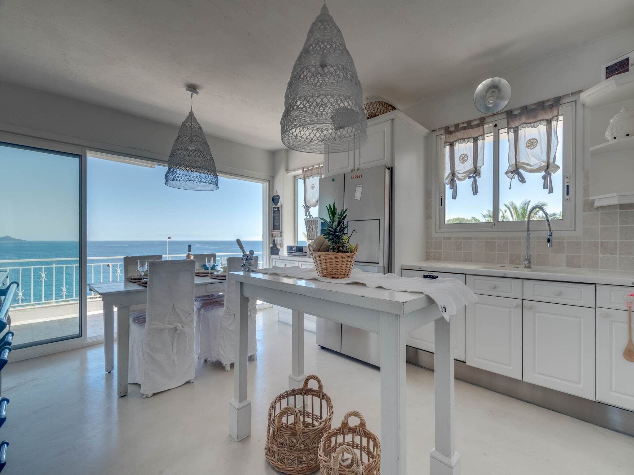 Een ruime keuken met witte kasten, een eethoek met een tafel voor twee en grote ramen met uitzicht op zee.
