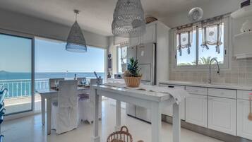 Een ruime keuken met witte kasten, een eethoek met een tafel voor twee en grote ramen met uitzicht op zee.