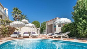 A swimming pool with lounge chairs and umbrellas, surrounded by greenery and a building with a tiled roof. - Villa in Les Issambres near the Sea (Les Issambres)