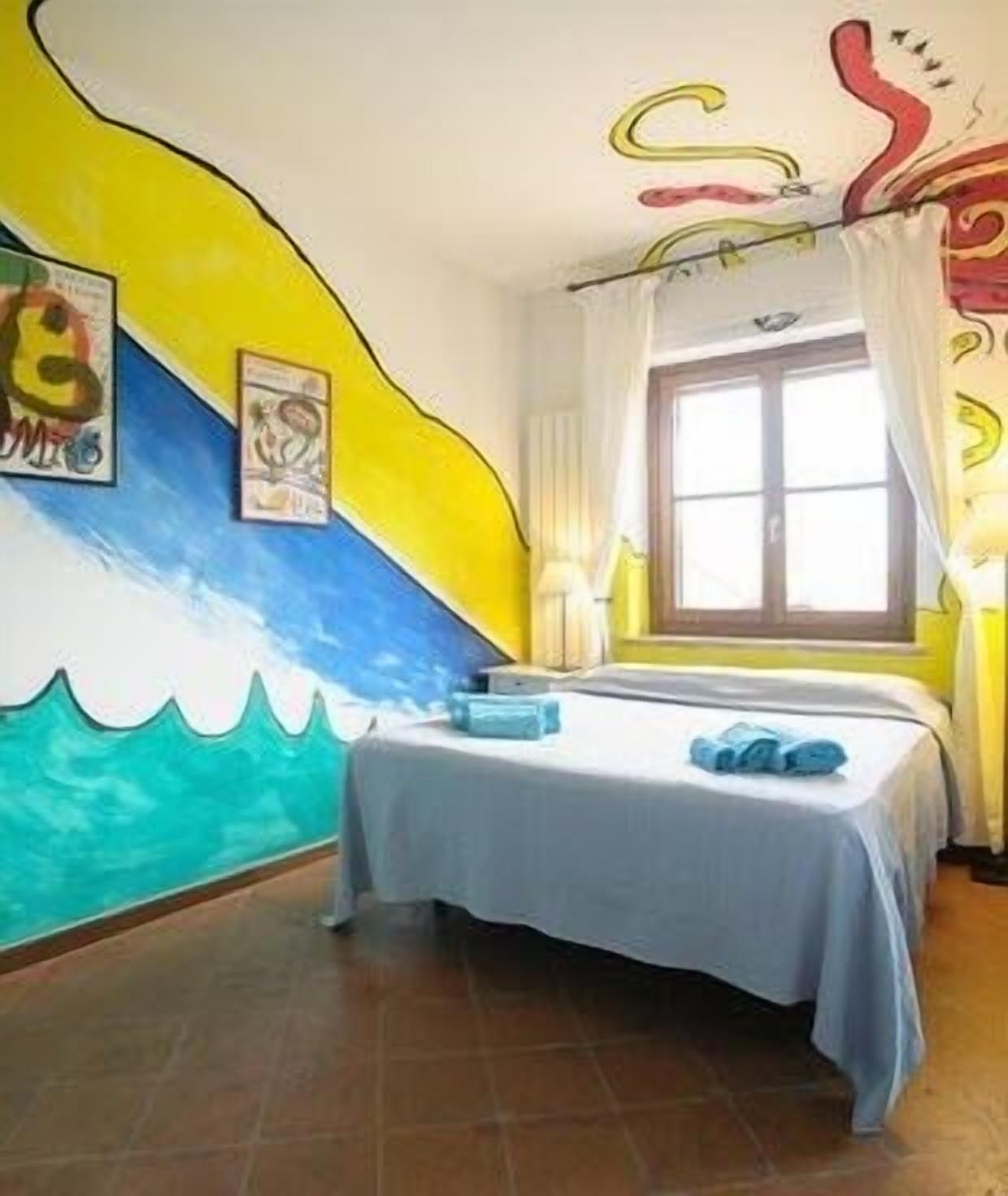 Une pièce avec un lit, une fenêtre avec des rideaux et un mur orné d’une fresque colorée.