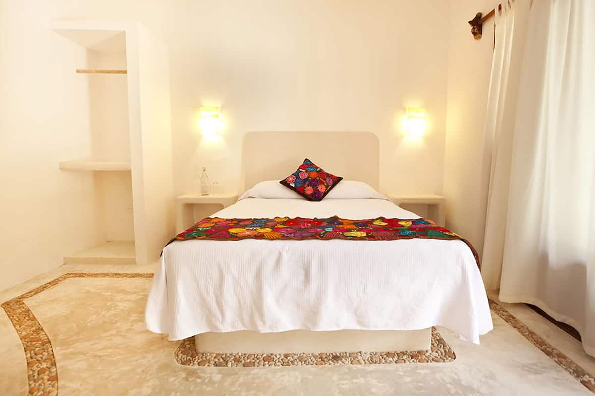 A bed with a white bedspread and a colorful blanket, two bedside lamps, and a nightstand with a bottle.