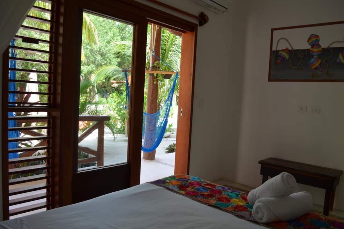A hotel room with a bed, a small table, a hammock, and a view of the outdoors.