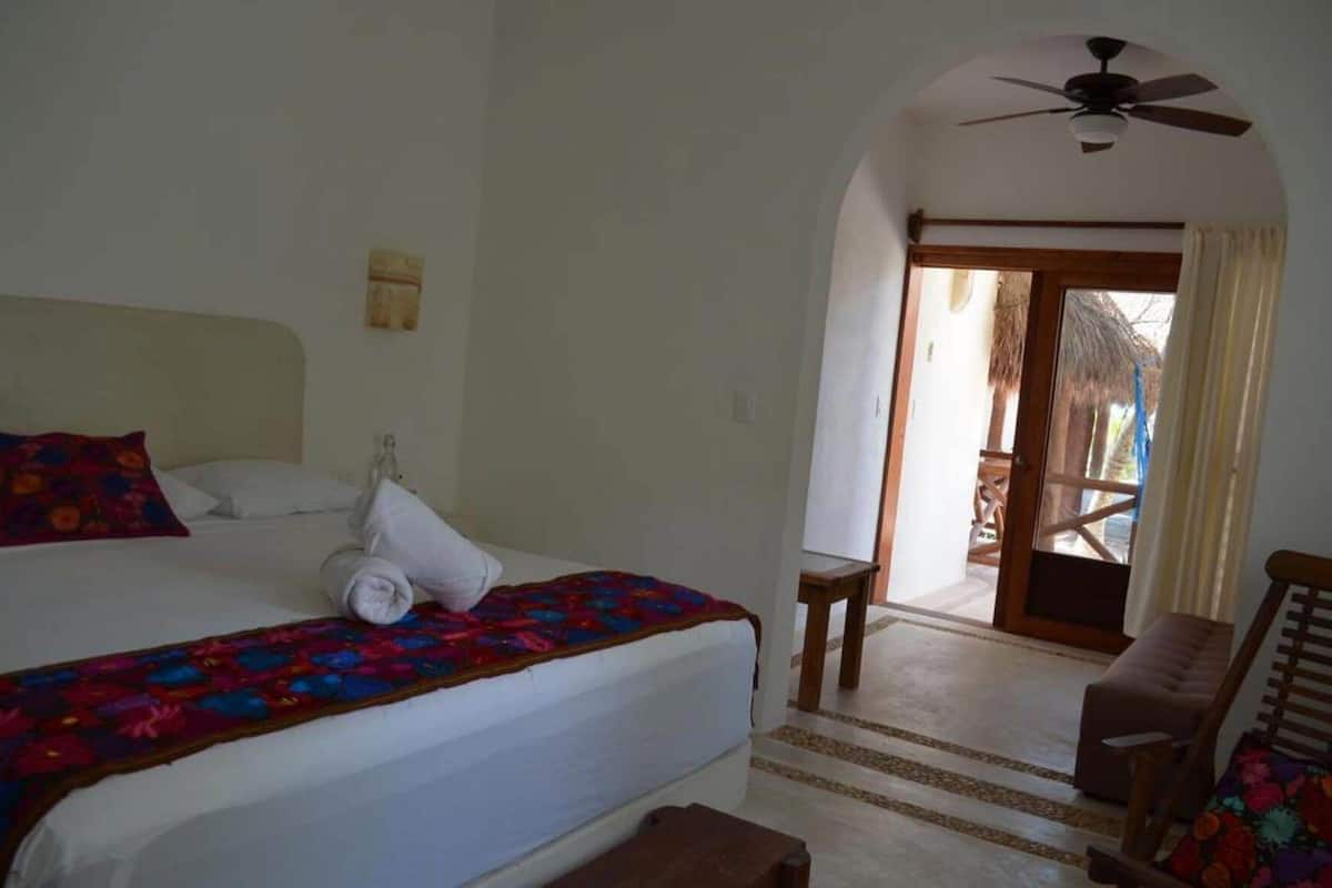 A hotel room with a bed, a chair, a small table, a ceiling fan, and a view of the outside.