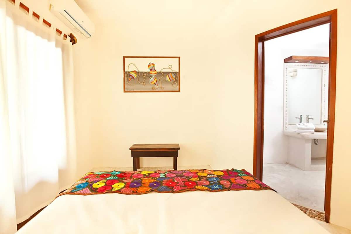 A bedroom with a bed, a small wooden table, a colorful quilt, a framed picture on the wall, and a bathroom visible through an open door.