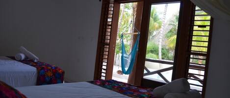 A room with two beds, a hammock, and a view of a balcony with palm trees.