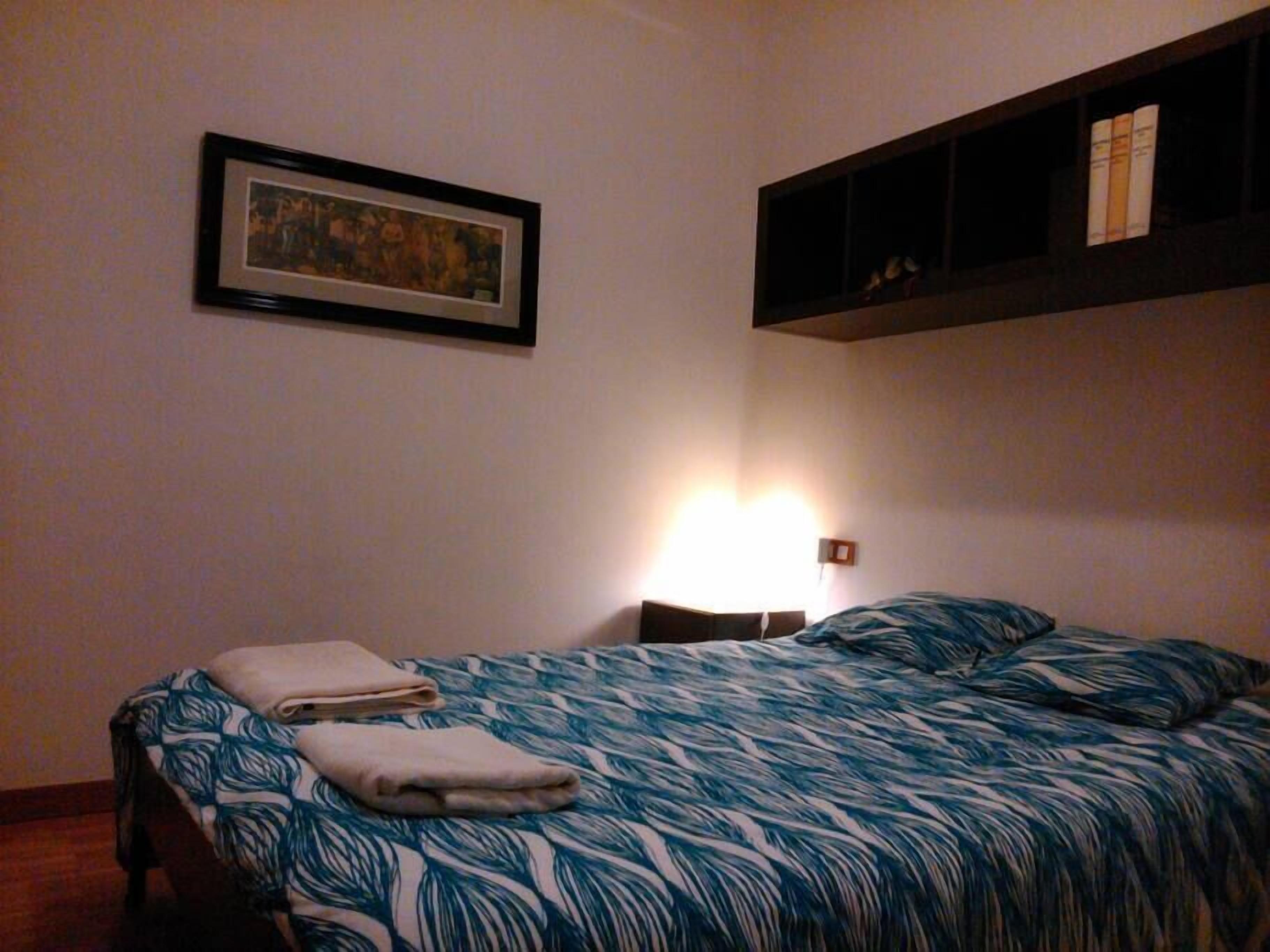 A bedroom with a bed, a framed picture on the wall, and a shelf with books.