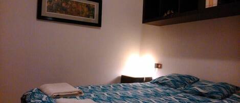 A bedroom with a bed, a framed picture on the wall, and a shelf with books.