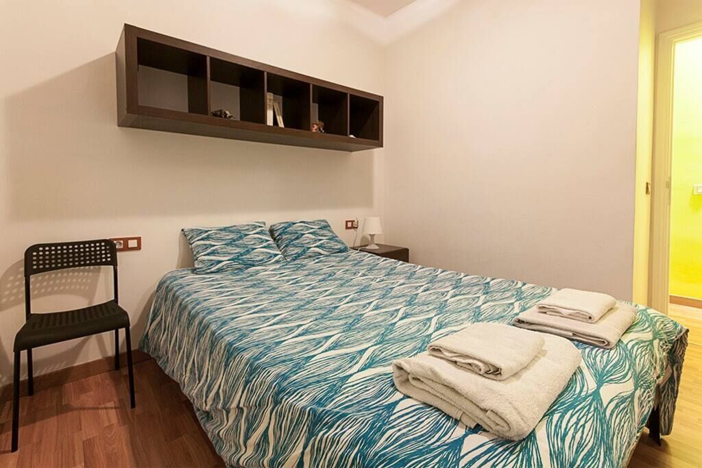 A bedroom with a bed, a chair, a nightstand, and a shelf above the bed.