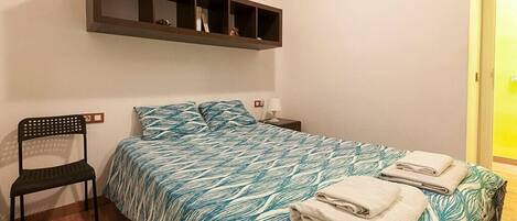 A bedroom with a bed, a chair, a nightstand, and a shelf above the bed.