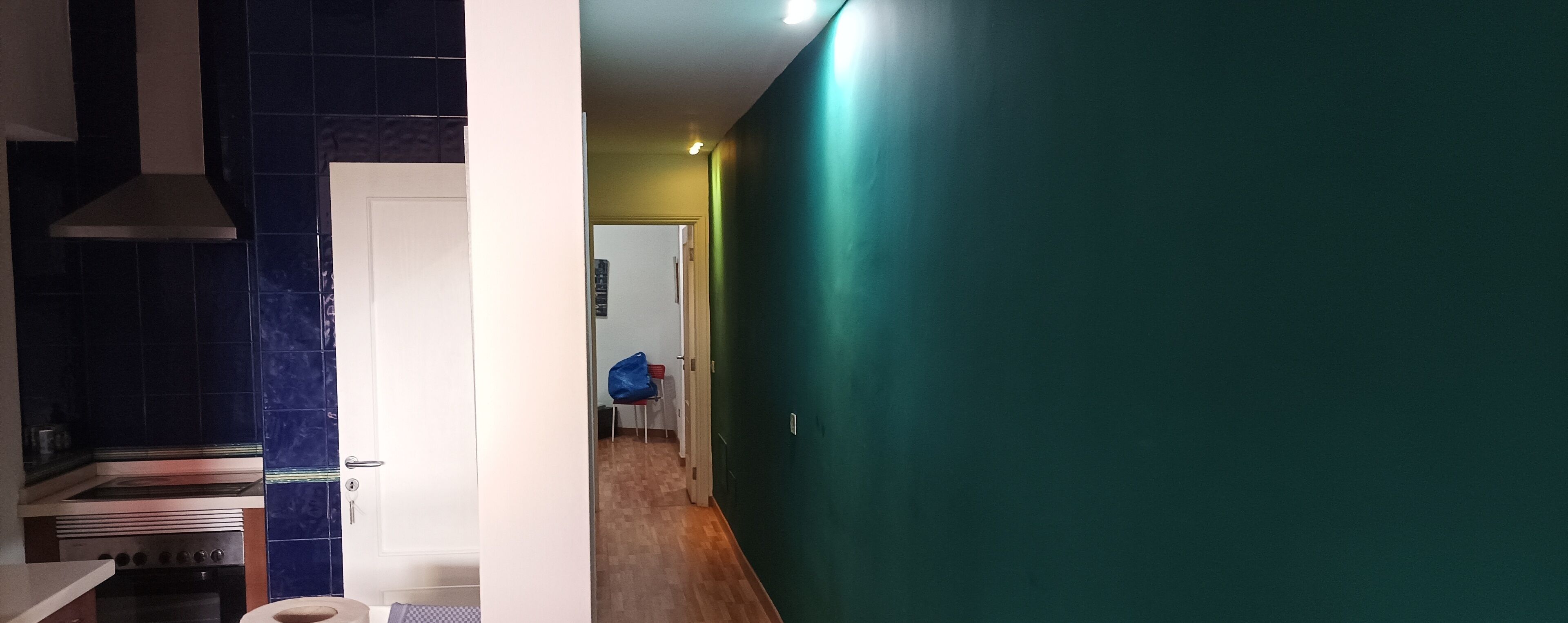 A kitchen with a stove, oven, and blue-tiled backsplash, leading to a hallway with a green wall and a door.