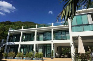 A modern multi-story hotel with large windows, outdoor seating, and a view of a lush green hillside.