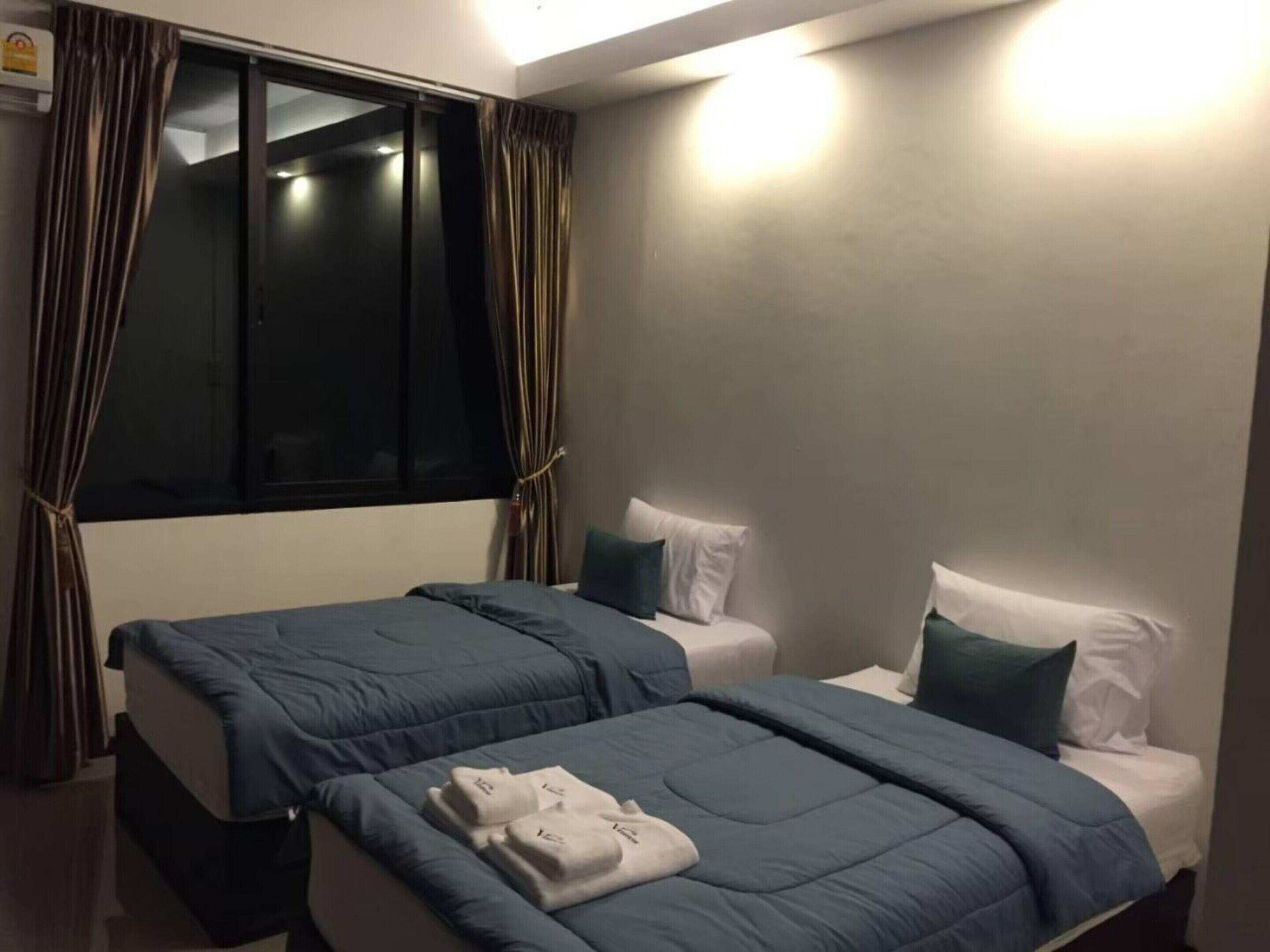 A hotel room with two beds, each with a blue bedspread and white pillows. There is a window with curtains, and a small table with a lamp.