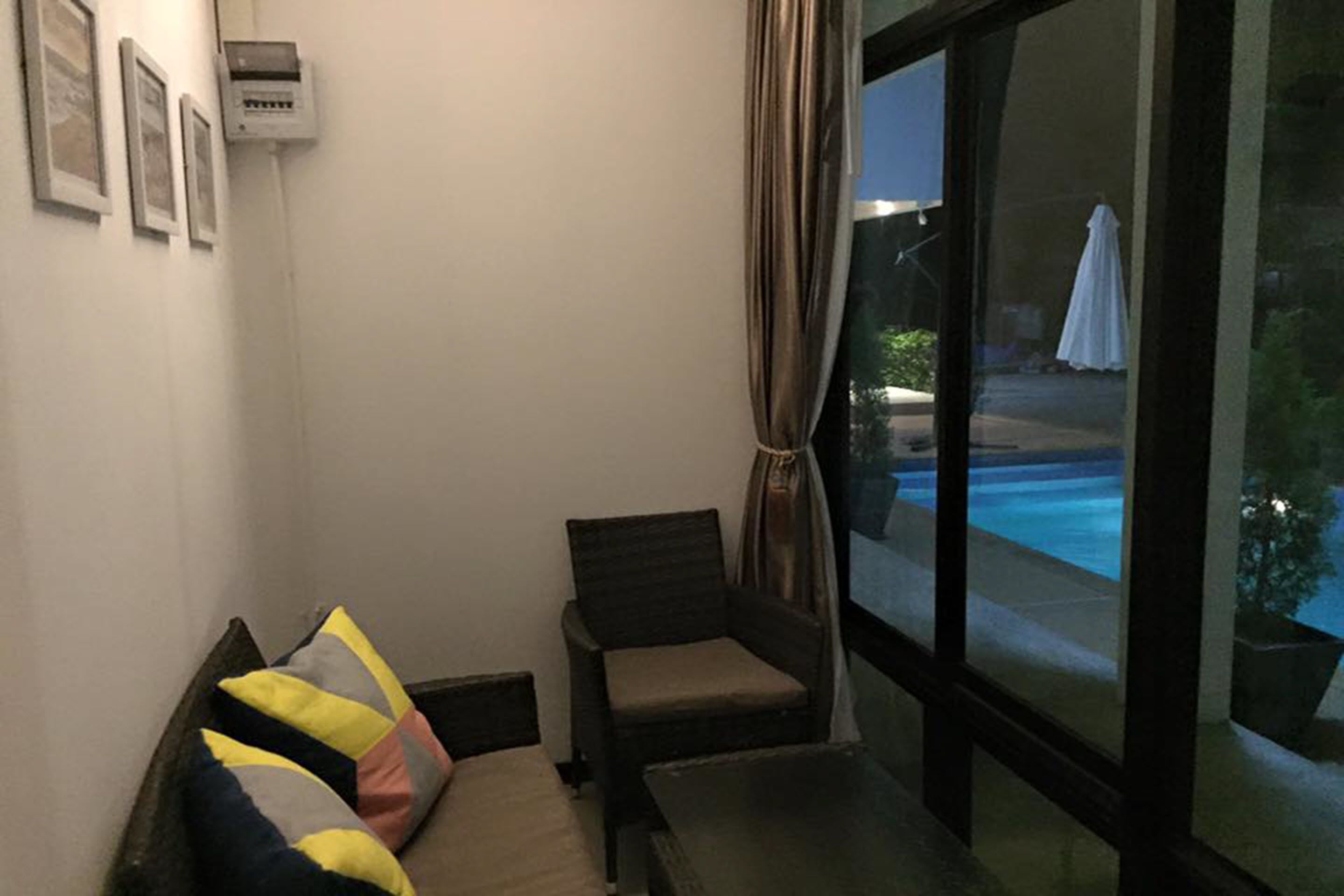 A room with a sofa, a chair, a window with a view of a pool and outdoor area, and a wall-mounted air conditioning unit.