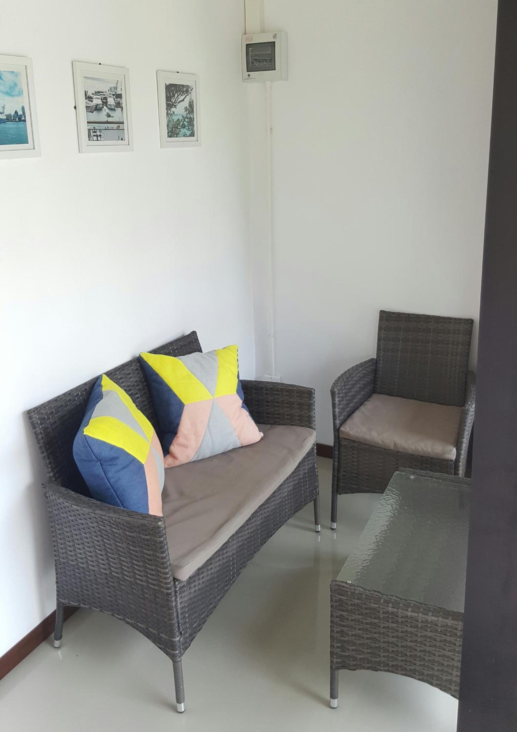 A room with a grey wicker sofa and armchair, a glass coffee table, and three framed pictures on the wall.