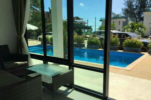 A room with a large window overlooking a swimming pool, wicker furniture, and a view of the street and buildings outside.