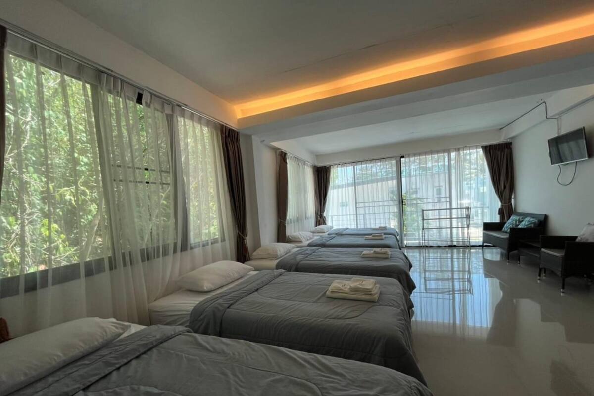 Une chambre d’hôtel avec trois lits, de grandes fenêtres avec des rideaux légers, un téléviseur à écran plat fixé au mur et un design épuré et moderne.