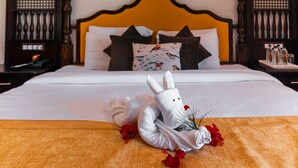 Una cama bien hecha con una colcha blanca, un cojín decorativo con estampado de pájaros y una toalla blanca con forma de conejo y flores rojas.