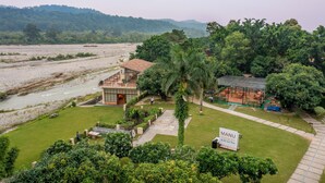 Un hotel con vista a un río y colinas, un área de comedor al aire libre techada, y un letrero que dice "MANU Marani".