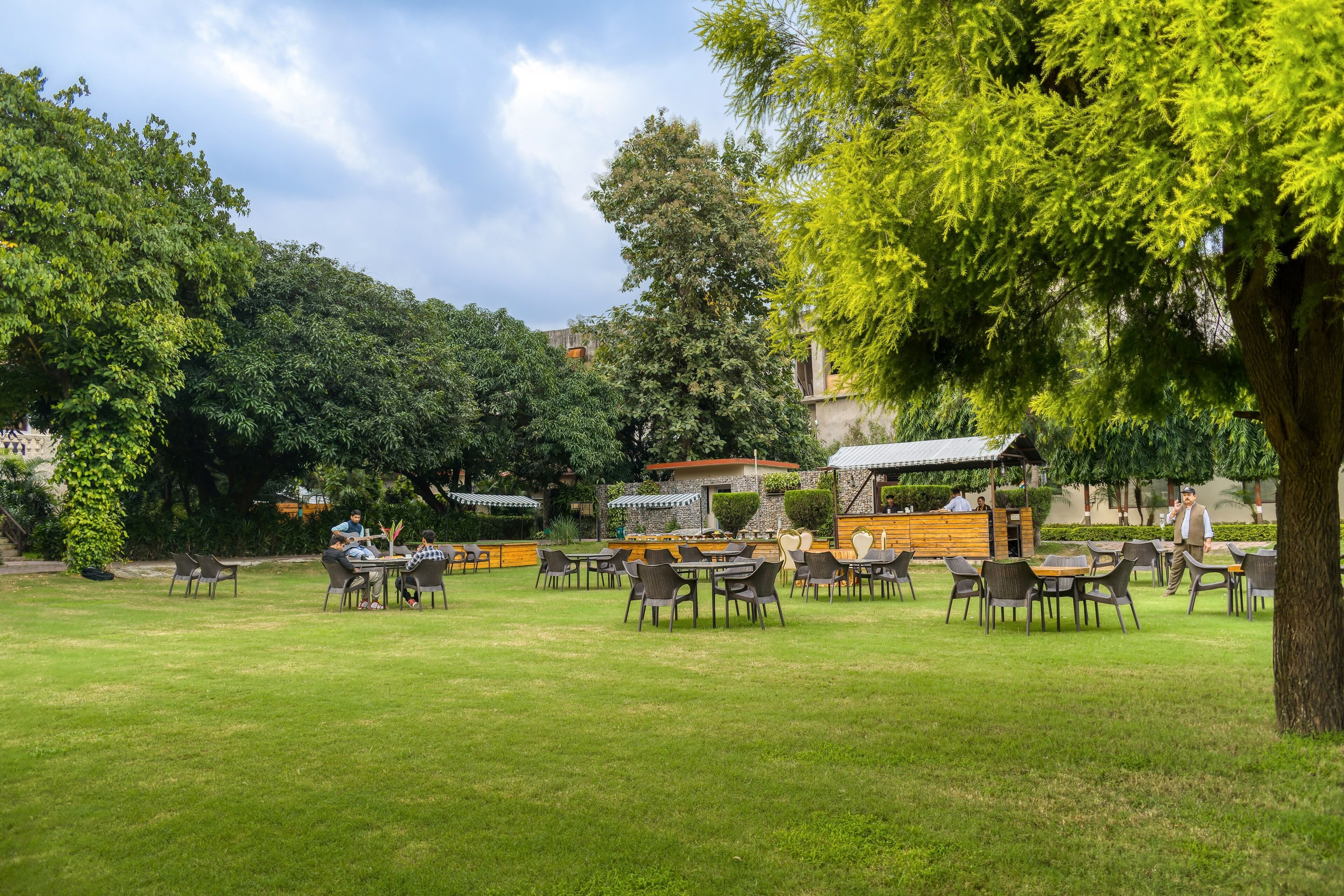 An outdoor seating area with tables and chairs, surrounded by trees and a building with a sloped roof.