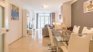 Ein moderner Essbereich mit einem Glastisch und weißen Stühlen, einem Sofa im Hintergrund und Kunstwerken an den Wänden.