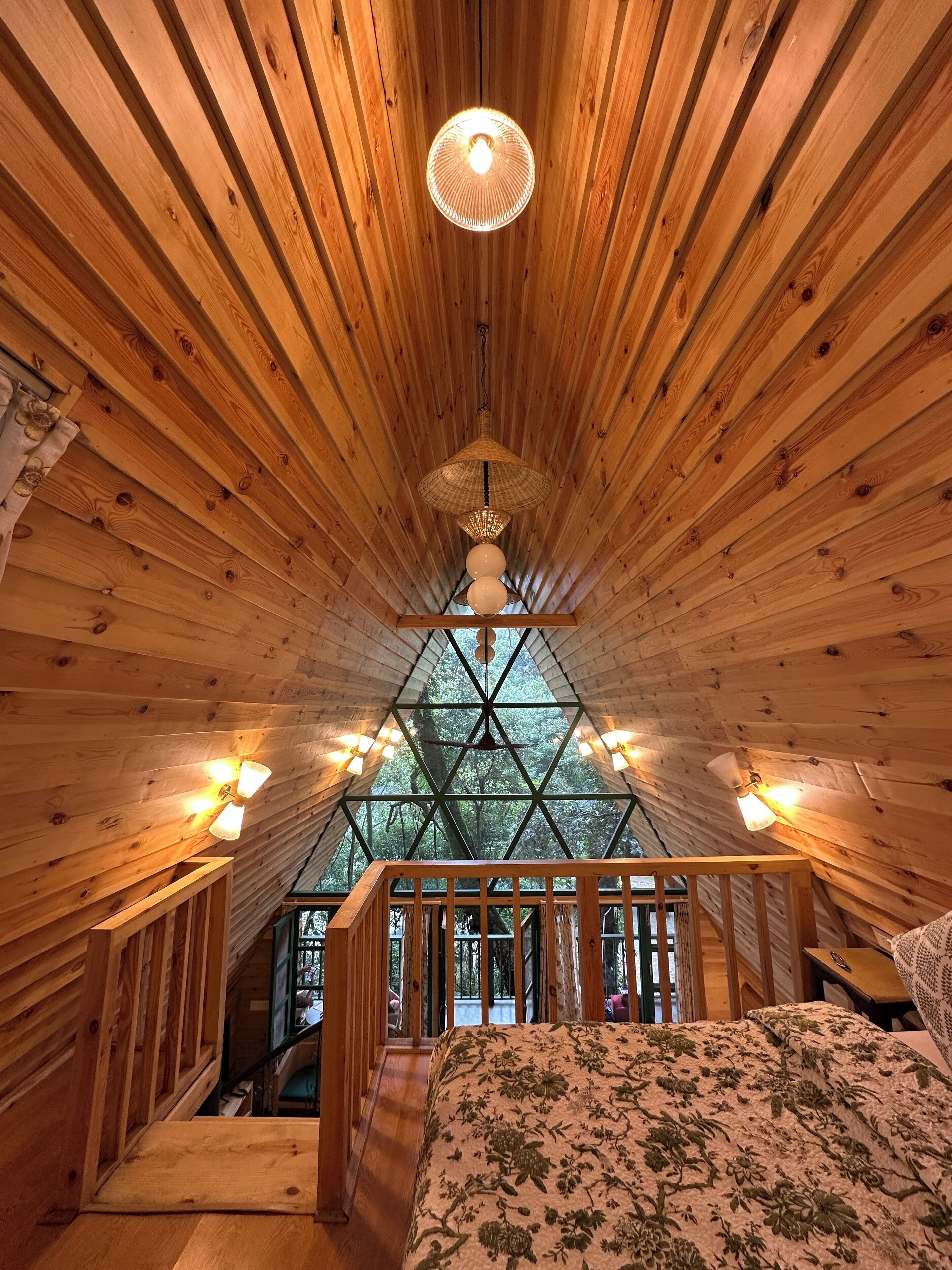 A wooden cabin interior with a sloped ceiling, a bed with floral bedding, and a view through a triangular window.