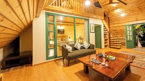 Um quarto acolhedor no estilo loft, com teto de madeira, janela com moldura verde, sofá, mesa de madeira e escada para um andar superior.