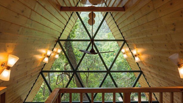 Uma cabana de madeira com design de janela triangular, uma cama com colcha florida e uma varanda com guarda-corpo.