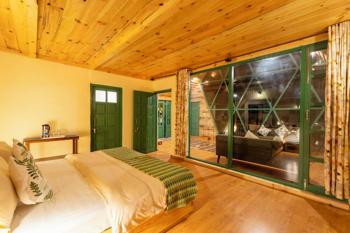 Een houten plafond, een bed met een groene deken, een groene deur en uitzicht door een glazen deur.