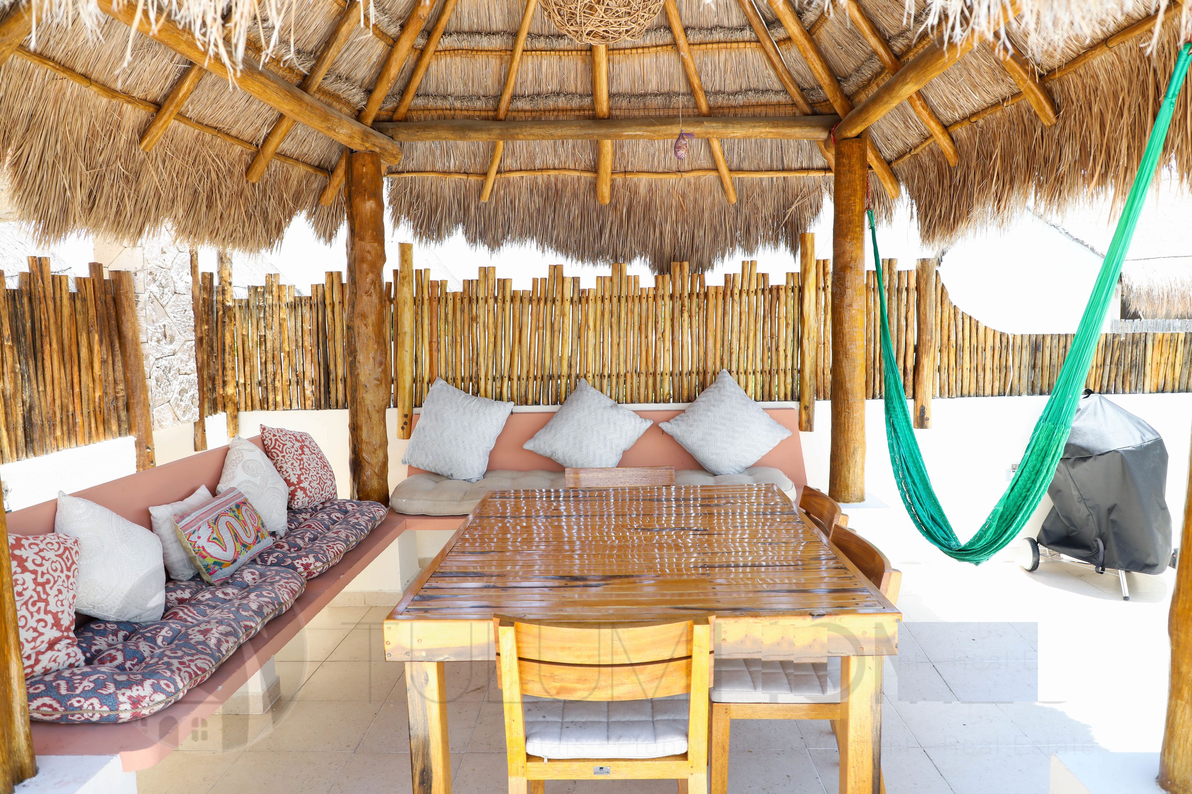 A wooden gazebo with a thatched roof, a cushioned bench, and a table with chairs.