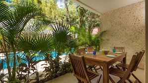 A wooden table with chairs, surrounded by lush green plants and a view of a swimming pool.