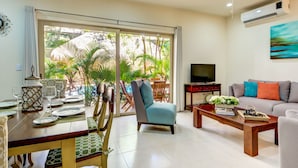 A living room with a dining area, a sofa, a TV, and a view of a garden through a sliding door.